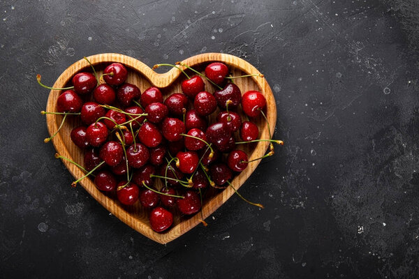 Ripe cherries and strawberries on a wooden plate in the shape of a heart and dark gray background, Valentines card with fruit