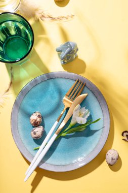  Paskalyayı kutlamak için masa ayarlayın. Tabak, çatal bıçak, yumurta ve bahar çiçekleri sarı masa üst görünümü, dikey fotoğraf