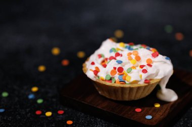 Homemade puff pastry tartlet with whipped cream, sprinkled with colored pastry sprinkles on a dark background.