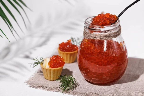 Red caviar in a glass jar with a teaspoon. Homemade tartlets made of puff pastry with red caviar and butter. On a white background, the shadow of fern leaves.