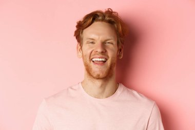 Dağınık kızıl saçlı, sakallı, gülüp gülümseyen, beyaz dişlerini gösteren pembe arka planda mutlu bir genç adam.