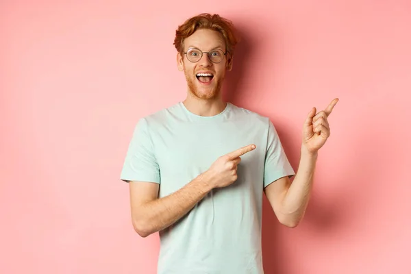Hombre pelirrojo sorprendido en gafas y camiseta mostrando promoción especial, señalando los dedos en la esquina superior derecha y mirando fijamente emocionado a la cámara, de pie sobre el fondo rosa — Foto de Stock