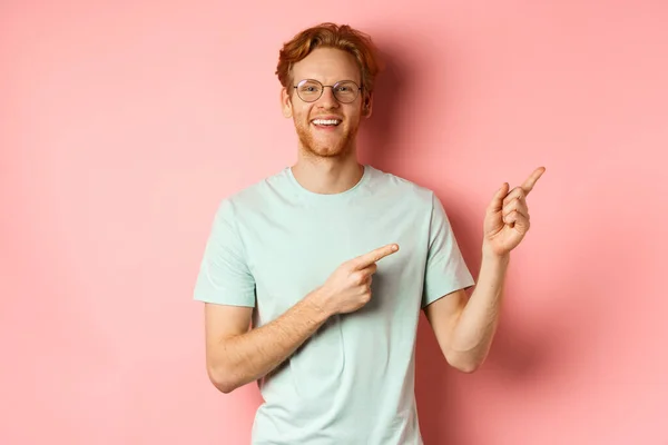 Amistoso pelirrojo con gafas apuntando a la esquina superior derecha, mostrando pancarta promocional y sonriendo a la cámara, de pie sobre fondo rosa — Foto de Stock