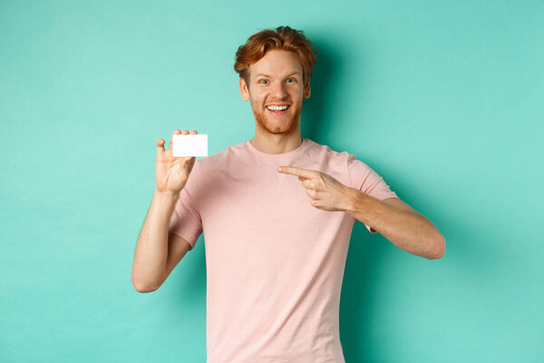 Attractive adult man with beard and red hair pointing finger at plastic credit card, smiling pleased at camera, standing over turquoise background