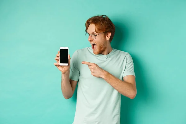 看看这个。戴眼镜的红头发帅哥用手指指着空白的智能手机屏幕，在网上显示促销效果，对蓝绿色的背景感到惊讶 — 图库照片