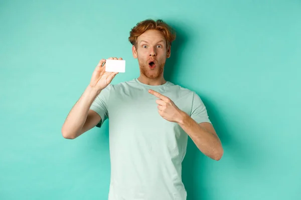 Koncepcja zakupów. Przystojny rudzielec w t-shircie z plastikową kartą kredytową i uśmiechnięty, stojący na turkusowym tle — Zdjęcie stockowe