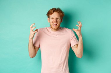 Annoyed and pissed-off redhead guy looking angry, shouting and grimacing, shaking hands aggressive, standing furious against mint background