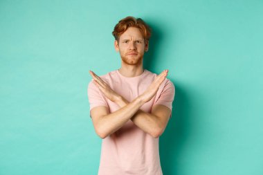 Tişörtlü kuşkucu kızıl adam hayır diyor, kollarını göğsüne dayayıp hayal kırıklığına uğruyor, kötü bir şeyi yasaklıyor, turkuaz arka planda duruyor.