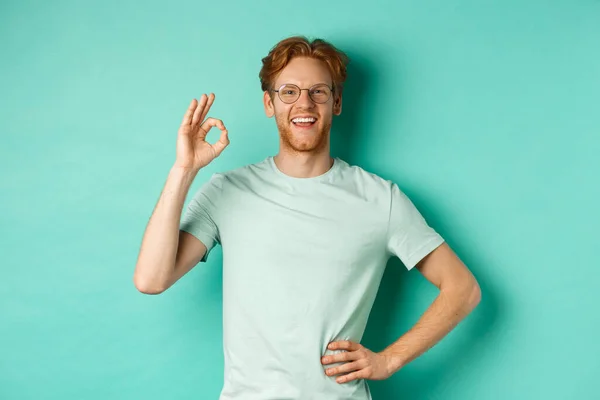 Joven feliz con el pelo rojo y la barba, con gafas y camiseta, sonriendo satisfecho y mostrando signo ok, decir sí, aprobar y estar de acuerdo, de pie sobre fondo turquesa — Foto de Stock