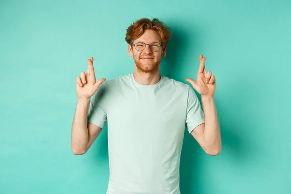 Joven chico alegre con el pelo rojo y la barba, con gafas, sonriendo y los dedos cruzados para la buena suerte, pidiendo deseos y buscando optimista, de pie sobre fondo de menta — Foto de Stock