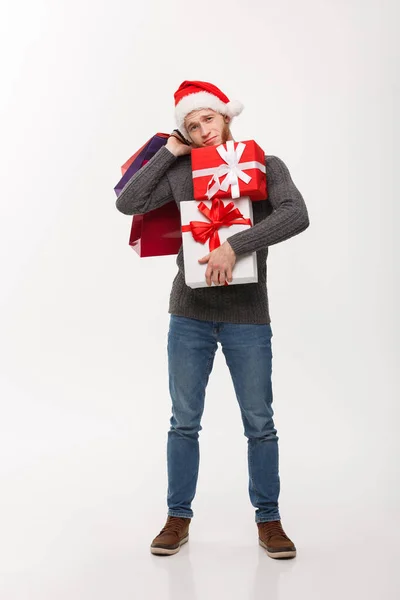 Boże Narodzenie koncepcja - młody, przystojny mężczyzna z broda trzymając ciężkie prezentuje i torby na zakupy z wyczerpany twarzy wyrażenie na białym tle. — Zdjęcie stockowe