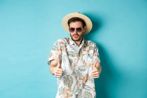 Turista bonito desfrutando de férias de verão, vestindo óculos de sol e camisa havaiana, mostrando polegar para cima na aprovação, de pé sobre fundo azul — Fotografia de Stock