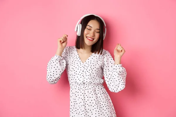 Feliz joven mujer asiática bailando y divirtiéndose, escuchando música en auriculares, de pie sobre fondo rosa — Foto de Stock