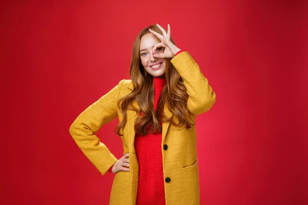 Menina brilhante e ensolarado sentindo-se feliz, mesmo em dias chuvosos de pé em amarelo casaco elegante sobre fundo vermelho mostrando gesto ok sobre os olhos, sorrindo amplamente para a câmera, segurando a mão na cintura — Fotografia de Stock