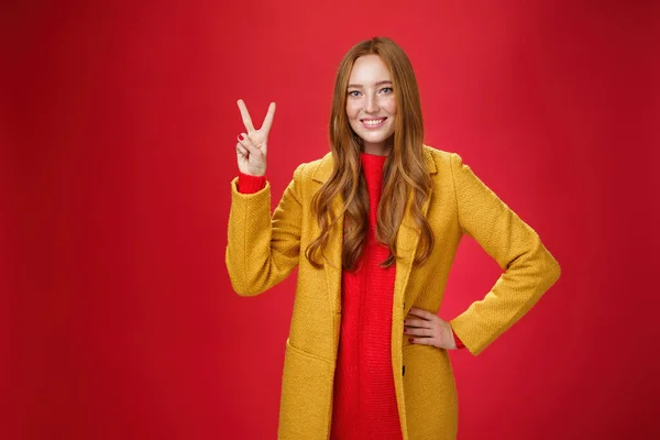 Portret van vrolijke speelse en schattige roodharige vrouw met sproeten in gele jas die hand in hand op taille vertrouwen en zelfverzekerd tonen vrede of twee teken blij, verslaan weersomstandigheden — Stockfoto
