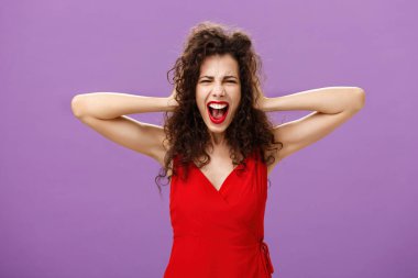 Ex-boyfriend spoilt woman formal event being outraged and pissed yelling in fury holding hands on curly hair grimacing from anger standing over purple background in stylish evening red dress