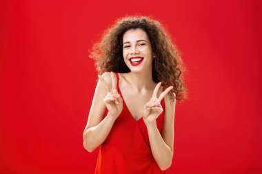 Arkadaş canlısı, sosyal, güzel, kıvırcık saçlı, kırmızı elbiseli ve akşam makyajlı, barış ve zafer işaretlerini gösteren mutlu bir kız.