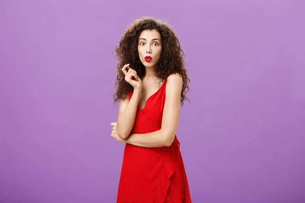 Заінтригована шахраюююча елегантна дама з кучерявою зачіскою в червоній сукні та губками, що розгинаються, дивно торкається волосини, яка вражена чутками, що поширюються навколо вечірки над фіолетовою стіною — стокове фото