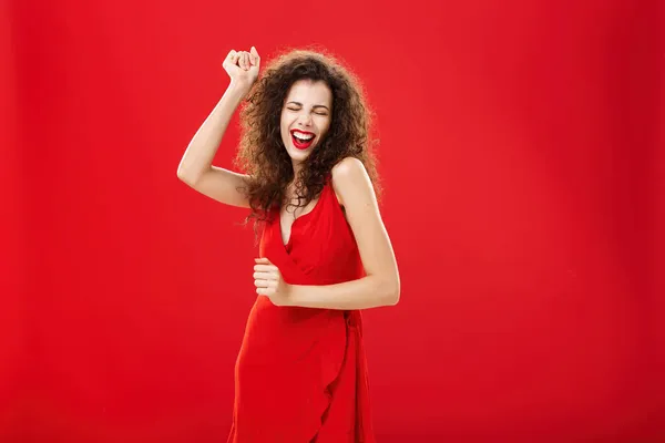 Πλούσια γυναίκα ξεχνά όλα τα προβλήματα στην πίστα. Χαριτωμένο κομψό νεαρό ευρωπαϊκό γυναικείο μοντέλο σε κομψό φόρεμα βράδυ κινείται στο ρυθμό της μουσικής πανηγυρισμούς ανύψωσης χέρι, τραγουδώντας θέτοντας πάνω από κόκκινο τοίχο — Φωτογραφία Αρχείου