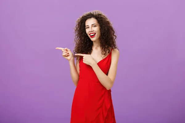 Mujer señalando a amigo presentando novio a padre indicando izquierda sonriendo ampliamente y mirando a la cámara despreocupada y amigable de pie en elegante vestido rojo sobre fondo púrpura usando maquillaje — Foto de Stock
