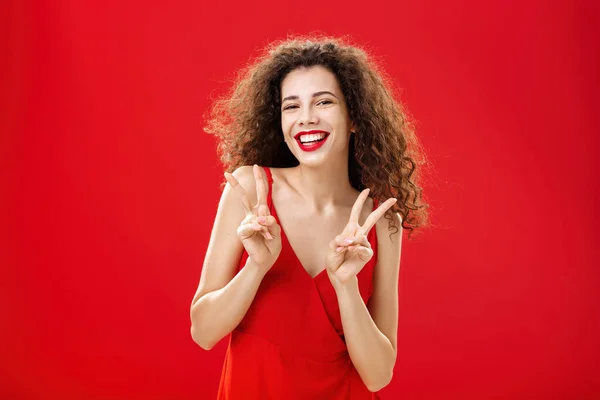 Φιλικό κοινωνικοποιημένο όμορφο χαρούμενο κορίτσι με σγουρό χτένισμα φορώντας κομψό κόκκινο φόρεμα και βραδινό make-up που δείχνει την ειρήνη ή τη νίκη σημάδια χαρούμενα θέτοντας πάνω από στούντιο φόντο διασκεδάζεται — Φωτογραφία Αρχείου