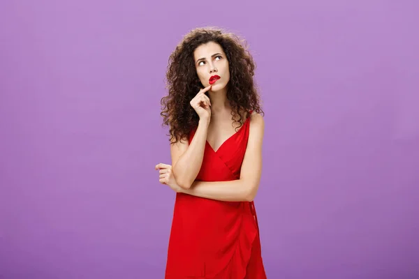 Elegante attente vrouw in trendy rode jurk met krullend kapsel en party make-up aanraken lip kijken naar de rechter bovenhoek denken hoe maken indruk tijdens gesprek over paarse achtergrond — Stockfoto