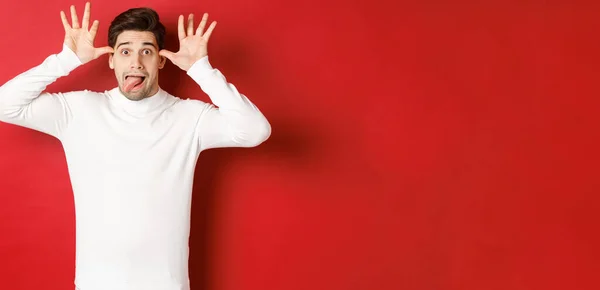 Portrait de gars caucasien drôle, montrant la langue et faire des visages, portant un pull blanc, debout sur fond rouge — Photo