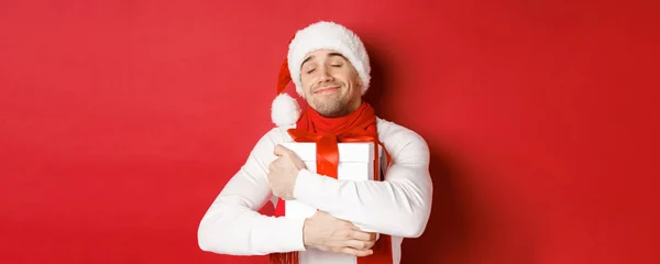 Concept of winter holidays, christmas and lifestyle. Image of lovely guy in santa hat and scarf, hugging his new year present and smiling flattered, standing over red background — Stock Photo, Image