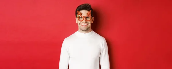 Gros plan de l'homme séduisant en pull blanc et lunettes de fête, célébrant Noël, souriant amusé, debout sur fond rouge — Photo