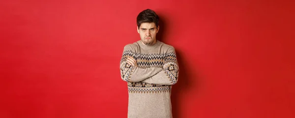 怒りと攻撃された男のイメージでクリスマスセーター,気が狂った感じ,胸と沈没のクロスアーム,赤の背景の上に立って — ストック写真