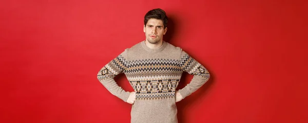 Portrait of doubtful and disappointed man, wearing christmas sweater, frowning and looking displeased at you, standing against red background — Stock Photo, Image