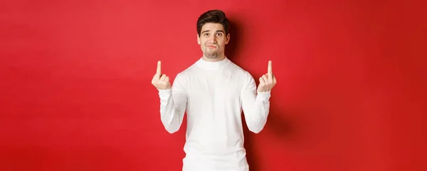 Image of pissed-off and distressed man telling to fuck off, showing middle-fingers and looking upset, standing over red background in white sweater — 스톡 사진