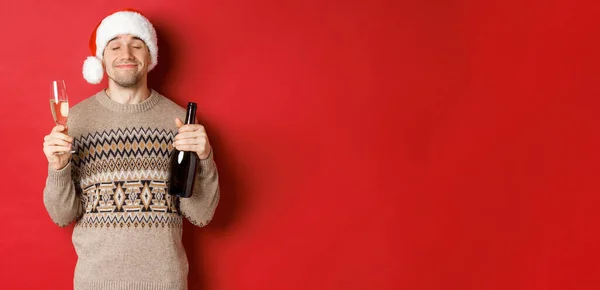 Conceito de férias de inverno, Natal e celebração. Imagem do homem sorridente satisfeito em chapéu de santa e suéter, bebendo no ano novo, segurando garrafa de champanhe e copo cheio, fundo vermelho — Fotografia de Stock