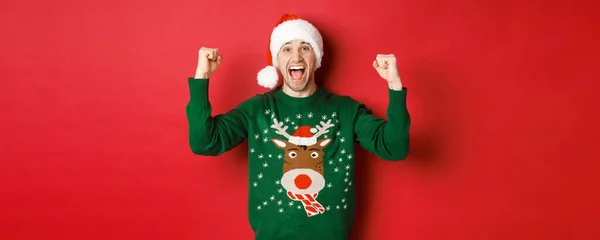 Retrato de homem atraente alegre comemorando o ano novo, vestindo suéter verde e chapéu de santa, gritando de alegria, ganhando ou triunfando, de pé sobre fundo vermelho — Fotografia de Stock