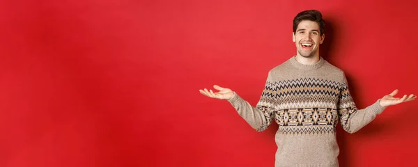 Portrait of happy good-looking man celebrating new year holidays, wearing christmas sweater, spread hands sideways and smiling, holding something on copy space, standing over red background — Stock Photo, Image