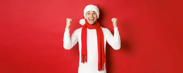 Noel Baba şapkası ve atkısı takmış mutlu ve heyecanlı bir adamın portresi, sevinen ve bir şeyler kazanan, yeni yılı kutlayan, kırmızı arka planda duran — Stok fotoğraf