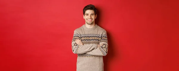크리스마스 스웨터를 입고 웃으며 카메라를 보고 크리스마스를 축하하며 빨간 배경 위에 서 있는 잘생긴 행복 한 남자의 사진 — 스톡 사진