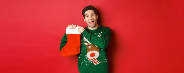 Imagem de homem alegre atraente em suéter, segurando meia de Natal com presentes, celebrando férias de inverno, de pé sobre fundo vermelho — Fotografia de Stock