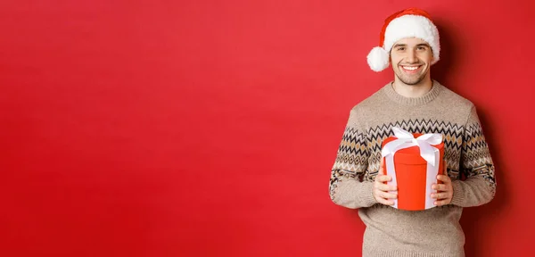 Portret van een knappe man die een cadeautje vasthoudt, een fijne vakantie wenst, in kerstmuts en kersttrui staat tegen een rode achtergrond — Stockfoto