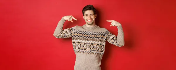 Concepto de celebración de Navidad, vacaciones de invierno y estilo de vida. Imagen de hombre guapo feliz en jersey de Navidad apuntándose a sí mismo y sonriendo, siendo santa secreta, de pie sobre fondo rojo —  Fotos de Stock