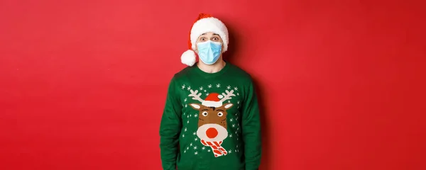 Concept van nieuwjaar, covid-19 en sociale afstand. Verrast man in santa hoed, medisch masker en kerst trui kijken verbaasd op camera, staande over rode achtergrond — Stockfoto