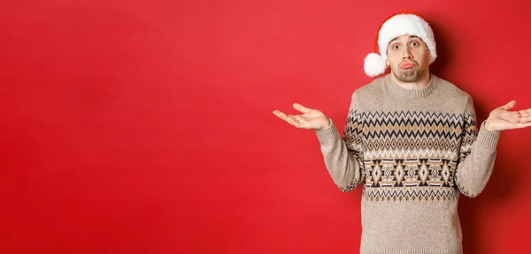 Imagem de jovem sem noção em suéter e chapéu de santa, encolhendo os ombros e olhando sem saber, não sabe o que comprar para feriados de ano novo, de pé sobre fundo vermelho — Fotografia de Stock