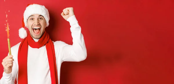 Концепция Рождества, зимних праздников и празднования. Красивый мужчина празднует Новый год и веселится, держа искру и улыбаясь, в шляпе Санты, стоя на красном фоне — стоковое фото