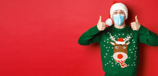 Conceito de ano novo, covid-19 e distanciamento social. Homem bonito alegre em chapéu de Papai Noel e máscara médica, mostrando polegares para cima, desfrutando de festa de Natal, de pé sobre fundo vermelho — Fotografia de Stock