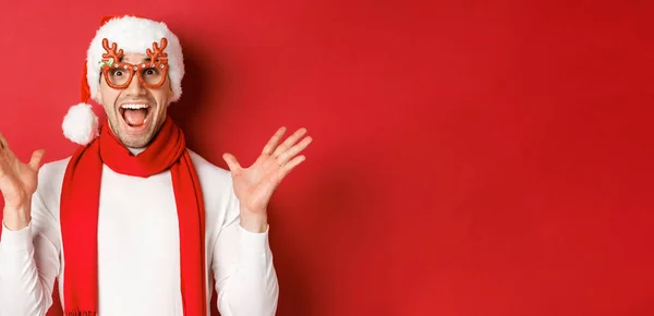 Έννοια των Χριστουγέννων, χειμερινές διακοπές και γιορτή. Εικόνα του έκπληκτος και χαρούμενος άνθρωπος που αναζητούν έκπληκτος, φορώντας γυαλιά κόμμα και απολαμβάνοντας το νέο έτος, στέκεται πάνω από το κόκκινο φόντο — Φωτογραφία Αρχείου