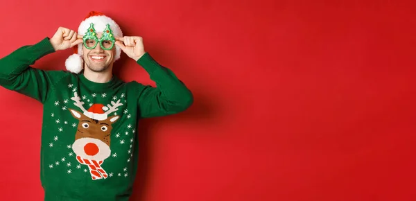 Портрет привабливого усміхненого чоловіка в капелюсі Санти, партійних окулярах і светрі, святкування новорічних свят, що стоять на червоному тлі — стокове фото