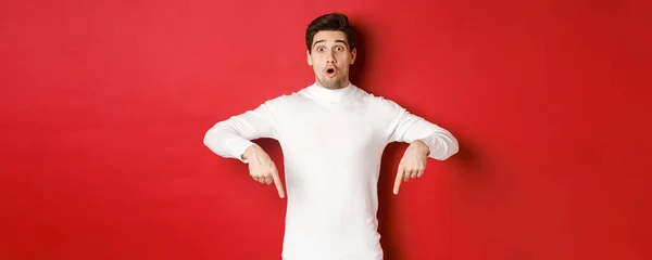 Изображение удивленного привлекательного парня в белом свитере, показывающего объявление, указывающего пальцами вниз и выглядящего изумленным, стоящего на красном фоне — стоковое фото