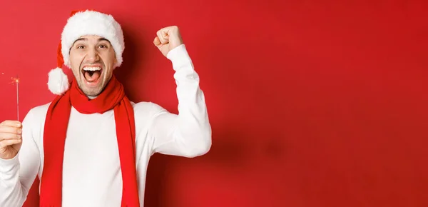 Концепция Рождества, зимних праздников и празднования. Портрет взволнованного красавца, поднимающего руку и держащего блеск, желающего счастливого нового года, стоящего на красном фоне — стоковое фото