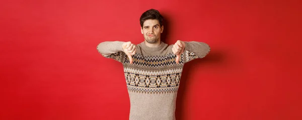 Portrait of skeptical and disappointed handsome man in christmas sweater, dislike new year party, showing thumbs-down, express disapproval, standing over red background — Stock Photo, Image