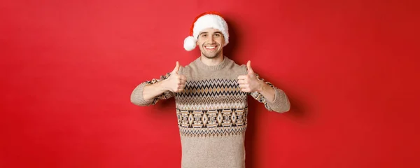 Image of cheerful attractive man in winter sweater and santa hat, showing thumbs-up, celebrating christmas and looking happy, like something, standing over red background — Stock Photo, Image